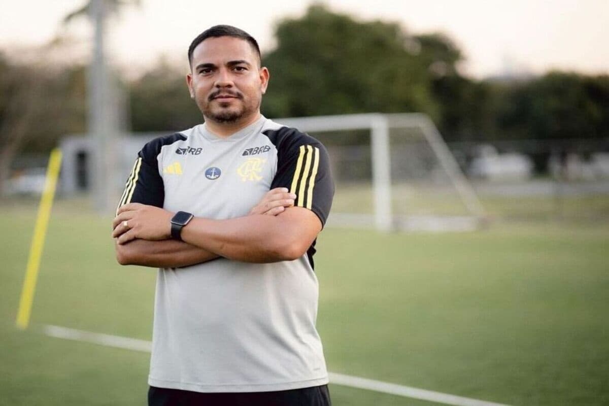 Filipe, técnico do sub-20 feminino está com o uniforme da comissão técnica, braços cruzados e ao fundo temos o campo da Gávea