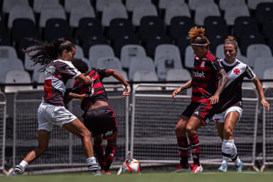 Valéria conduz a bola no clássico entre Flamengo e Vasco, enquanto uma jogadora adversária a segura pela camisa, Cristiane se aproxima para receber a bola e é marcada por outra atleta do Vasco. Ao fundo é possível ver a arquibancada de São Januário