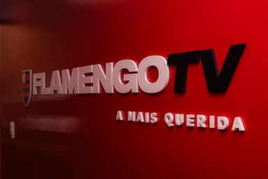 Logo da Flamengo TV com o escudo do Flamengo e o slogan “A Mais Querida” em parede vermelha.