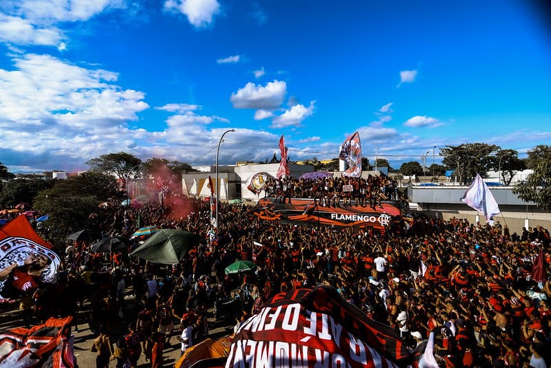 Multidão de torcedores do Flamengo realiza um grande AeroFla, com bandeiras, fumaça vermelha e o ônibus da delegação cercado por rubro-negros em festa sob céu azul.
