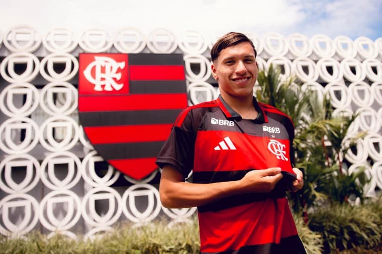 Juan Sayago posa com a camisa Flamengo em frente ao módulo da base no Ninho do Urubu