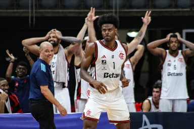 Gui Deodato sério após ponto contra Paulistano; ao fundo Oveja e companheiros celebram cesta do FlaBasquete